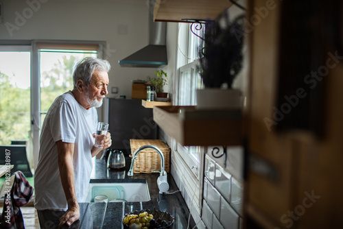 Senior man drinking water, thoughtful by window in home kitchen