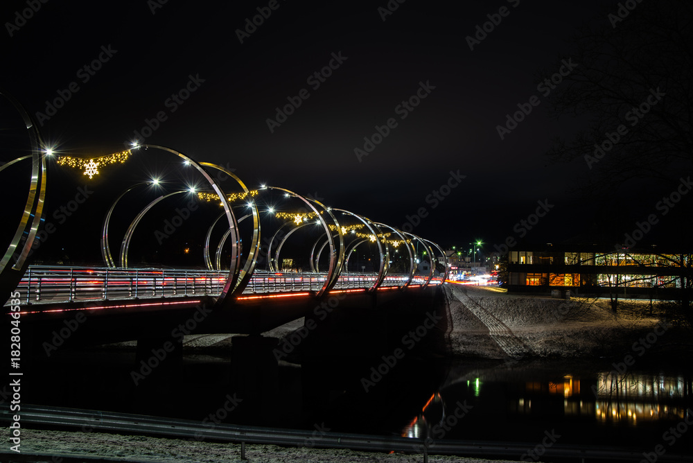 Obraz premium Night long exposure of a modern bridge with festive lights and traffic streaks over a dark river, winter banks and city glow.