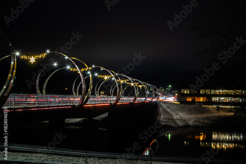 Night long exposure of a modern bridge with festive lights and traffic streaks over a dark river, winter banks and city glow.