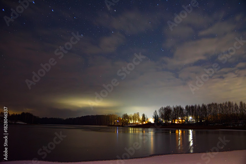 Starry winter night over a frozen lake, clouds drifting above silhouetted trees and distant lights reflecting on ice.