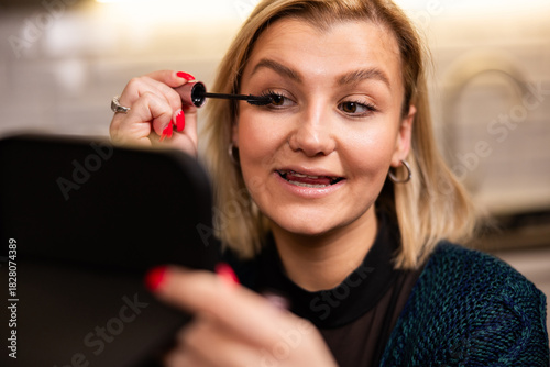 Woman applying mascara in a cozy kitchen setting while preparing for an evening out with friends