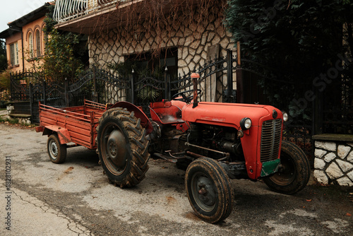 Fototapeta Naklejka Na Ścianę i Meble -  A red tractor with a small trailer stands on a quiet village road. The scene reflects rural work and agricultural lifestyle