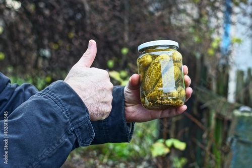 Fototapeta Naklejka Na Ścianę i Meble -  One glass small lidded jar with preserved green homemade traditional delicious pickled cucumbers in the hand of a man with a like during the day on the street