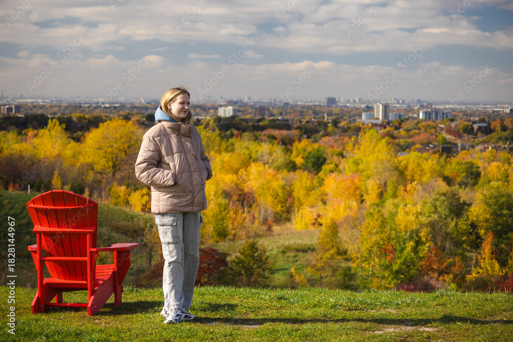 Fototapeta premium Young woman standing on a hill with hands in pockets, enjoying panoramic autumn views of Toronto skyline and colorful forest below