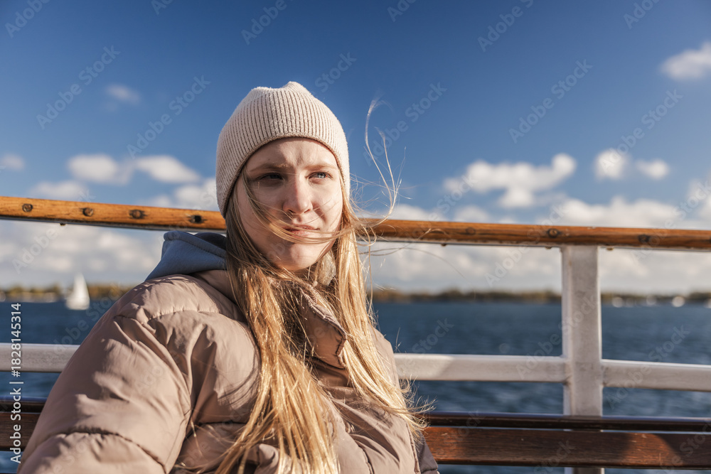 Obraz premium Young woman in a warm jacket and knit hat sitting on a boat on a sunny windy day over Lake Ontario in Toronto, Canada