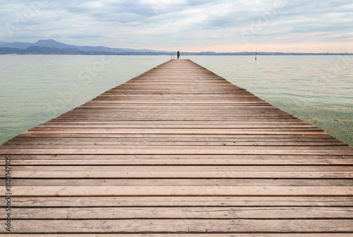 A long wooden pier extends into calm water under a soft, cloudy sky, with distant mountains on the horizon and a lone person standing at the end, creating a peaceful, minimalist scene.
