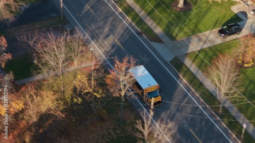 Aerial view of a yellow school bus driving along a quiet suburban street in a small American town surrounded by trees and residential houses.