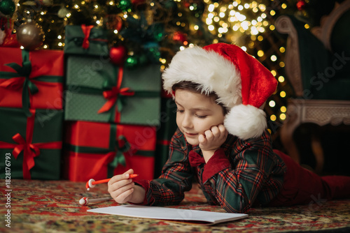 A child wearing a Santa hat and a plaid shirt with a bow tie writes a letter to Santa under the glowing Christmas tree lights. Pure holiday magic