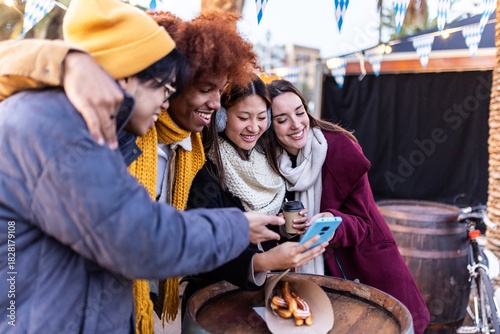 Group of multi-ethnic friends looking at photos on a smartphone at christmas market, hugging, smiling, and having fun together