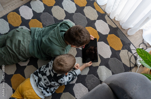 Top view of two brothers lying on colorful geometric carpet watching tablet together in living room at home. Concept of siblings digital bonding, screen time entertainment and modern childhood leisure