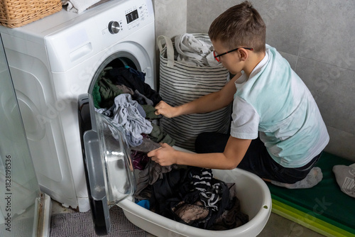 Boy with glasses unloading clean laundry from washing machine into plastic basin in bathroom at home. Concept of children helping with household chores, teaching responsibility and life skills early.