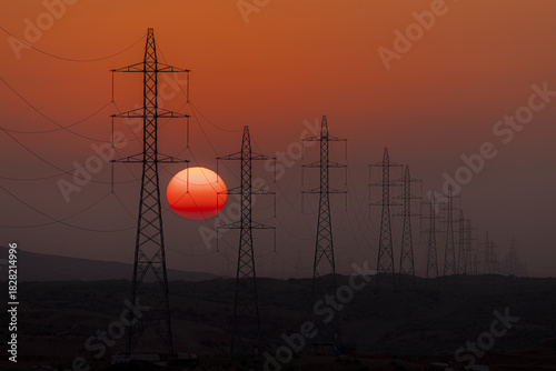 View of stark power lines cutting across the horizon as the sun sinks, painting the sky with fire, contrasting industrial presence with natural beauty, Rub' al-Khali, United Arab Emirates.