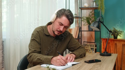 Middle-aged man at home listens to audiobook on smartphone headphones and writes notes focused on study. Young adult guy at table studies carefully absorbing knowledge writing key points for success