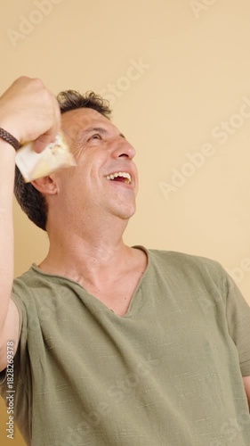 Middle-aged man with graying hair smelling and fanning his face with a stack of fifty euro banknotes, expressing greed and happiness. Vertical