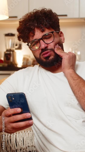 Arabian man at home feels tooth pain holds cheek opens smartphone search for dentist finds time. Young guy on sofa reads reviews picks dentist sends message books visit for pain relief needs care.