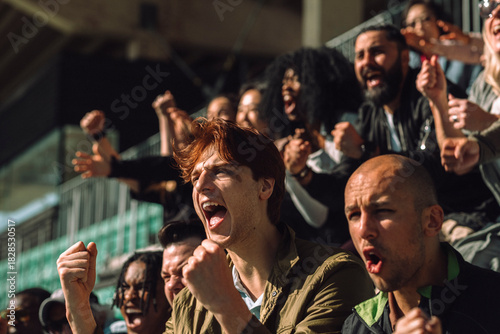 Excited young man shouting while cheering and making fist at sports stand on sunny day