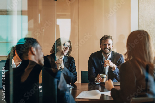 Smiling male and female lawyers sitting with colleagues in meeting room at office seen from glass