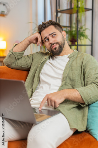 Overtired Caucasian young man with brown hair bored using laptop while sitting on sofa at home. Sleepy guy cannot concentrate on work and types lazily. Adult male is not interested in doing a project.