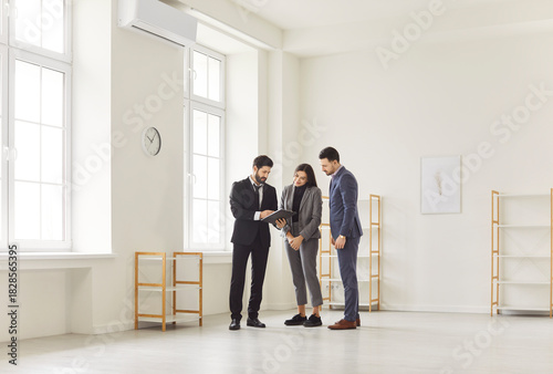 Real estate agent and couple clients in empty room, businesspeople standing, looking at tablet, discussing office plans, evaluating space, representing new property viewing or office planning