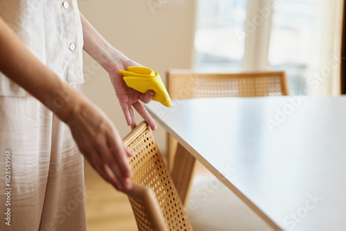 Cleaning woman wiping rattan chair and white table with a yellow sponge in a bright home interior, hands and arm visible, tidy living room scene showing domestic furniture care and routine.