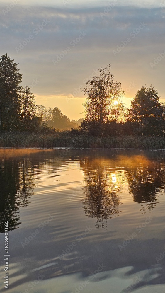 Fototapeta premium A boat ride on a lake at sunrise. Morning light. Tree silhouettes. Fog and reflection.