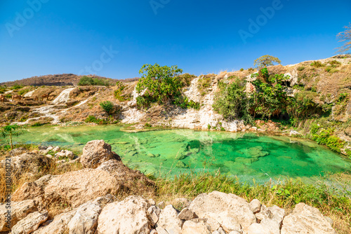 Wadi Darbat natural park in Dhofar, Oman, presenting clear emerald water with waterfalls cascading over lush rocks and green trees under a bright blue sky