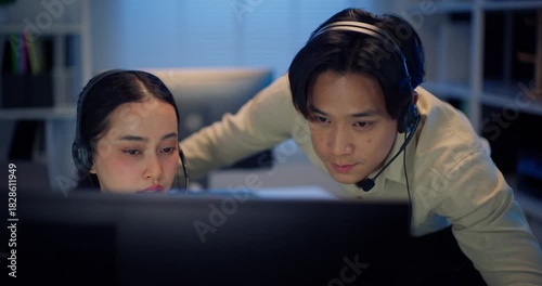 An Asian manager/supervisor assists a female call center agent while they both wear headsets, looking at a computer monitor in a dark office. Conveys support and training.
