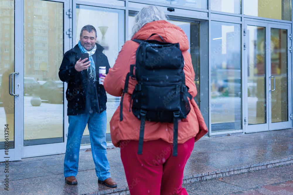Obraz premium Female photographer in red jacket filming man with phone and coffee cup in snowy weather outside modern business center. High quality photo
