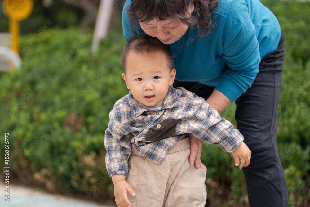 Naklejka premium An Asian grandmother tenderly adjusting the clothes of her 16-month-old grandson