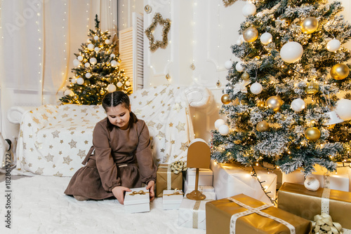 Little girl excitedly opens presents in a cozy holiday setting with festive decorations and sparkling lights