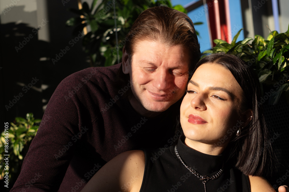 Obraz premium Middle-aged man and woman enjoying a moment together, with sunlight illuminating their faces, surrounded by lush green plants, conveying warmth and connection in a cozy atmosphere