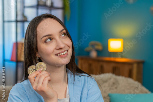 Young happy woman at home excitedly holding bitcoin BTC coin surprised by cryptocurrency profit, making wow gesture. Caucasian girl happily thinking about investments, analyzing market opportunities