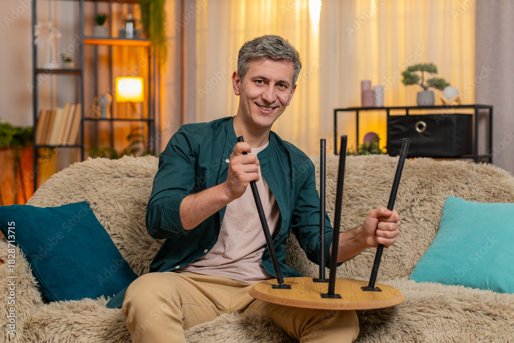 Fototapeta premium Young man at home repairs small wooden table with calm focus, appearing satisfied with progress and effort. Caucasian guy on sofa looks concentrated while enjoying handywork and fixing furniture part