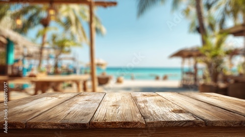 A wooden table sits in the foreground offering a view of a beautiful beach with palm trees and beach huts under a clear blue sky. The scene suggests relaxation and leisure.