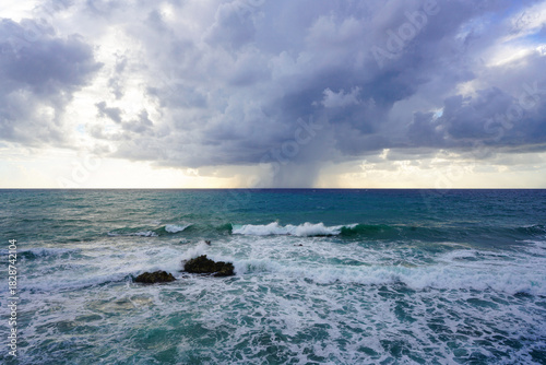 Dramatic view of strong wind and high waves crashing over rocks under a dark, brooding storm sky. Intense marine landscape with a visible rain squall on the horizon.