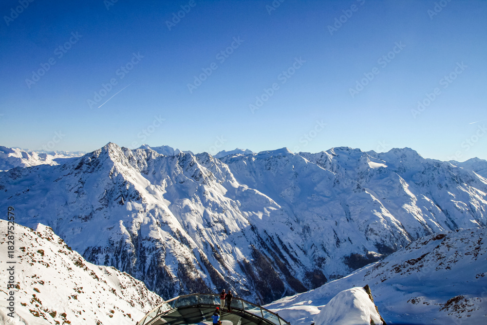 Naklejka premium A stunning panoramic view of the snow-capped Alps under a clear blue sky. A winter landscape with majestic peaks, icy slopes, and a glass observation deck overlooking the valley.