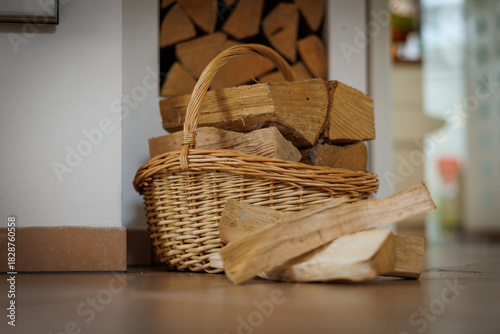 A cozy indoor scene featuring a wicker basket filled with neatly cut firewood placed beside a stack of logs.