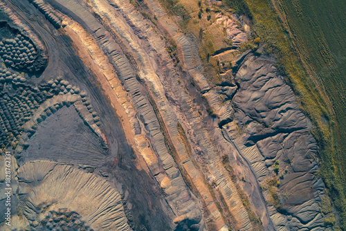 Top-down drone shot showing large, complex fan-shaped piles of dark industrial waste and exposed, layered geological terraces. Abstract texture of destructive impact of industrial extraction.