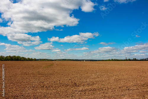 Vast plowed brown field stretches beneath a bright blue sky dotted with fluffy white cumulus clouds, promising fertile spring growth