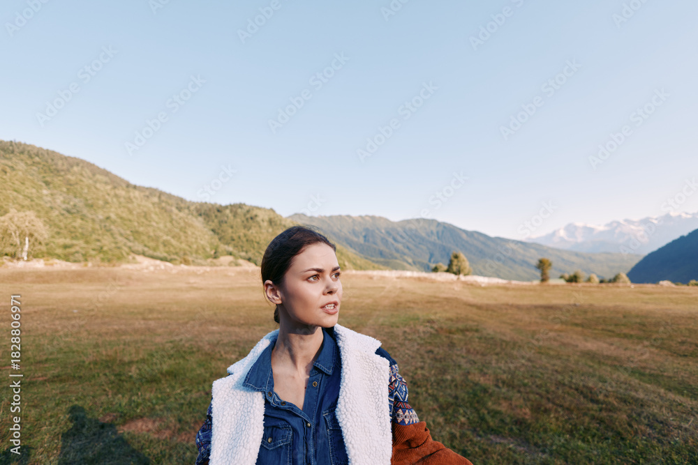 Fototapeta premium Woman portrait in meadow with mountains landscape, outdoor nature and travel mood. Young female in denim jacket and shearling coat gazing to the side across grassy field.
