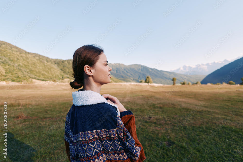 Fototapeta premium Woman in patterned jacket standing in meadow, portrait and profile view with mountains and landscape in background. Travel scene with serene nature, grass field and clear sky.