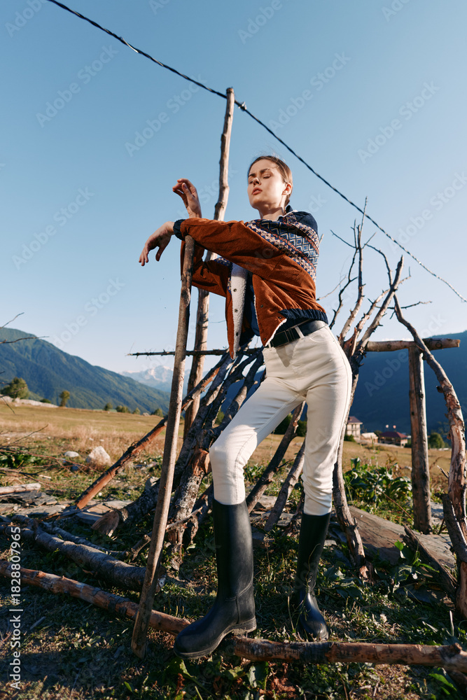 Fototapeta premium Woman model countryside mountains pose by rustic wooden structure wearing sweater, scarf and boots in field. Fashion portrait outdoor with natural landscape and casual style.