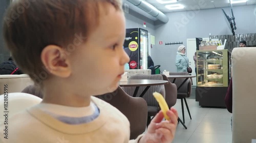 A cute little blonde baby sits in a cafe in a high chair and eats French fries, dessert, and drinks a cocktail. relaxes with her family. Happy childhood. Adorable little boy having breakfast at resort