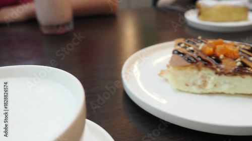  On the table of a cozy cafe there is a cup of aromatic cappuccino with creamy foam and a delicious dessert. A girl is eating it with a fork. In the background, people are looking at their phone and s