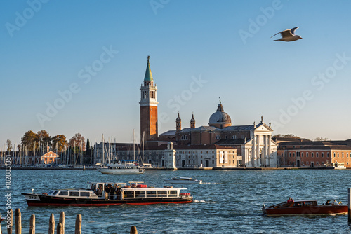 Venice lagoon view of San Giorgio Maggiore island with bell tower, basilica, vaporetto and boats crossing water while seagull soars overhead.