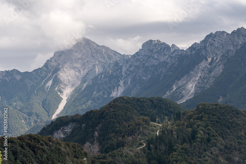 Dramatic Monte Sernio peak emerging from clouds above lush Friulian Alps forest in Friuli Venezia Giulia, Italy, under moody overcast sky.