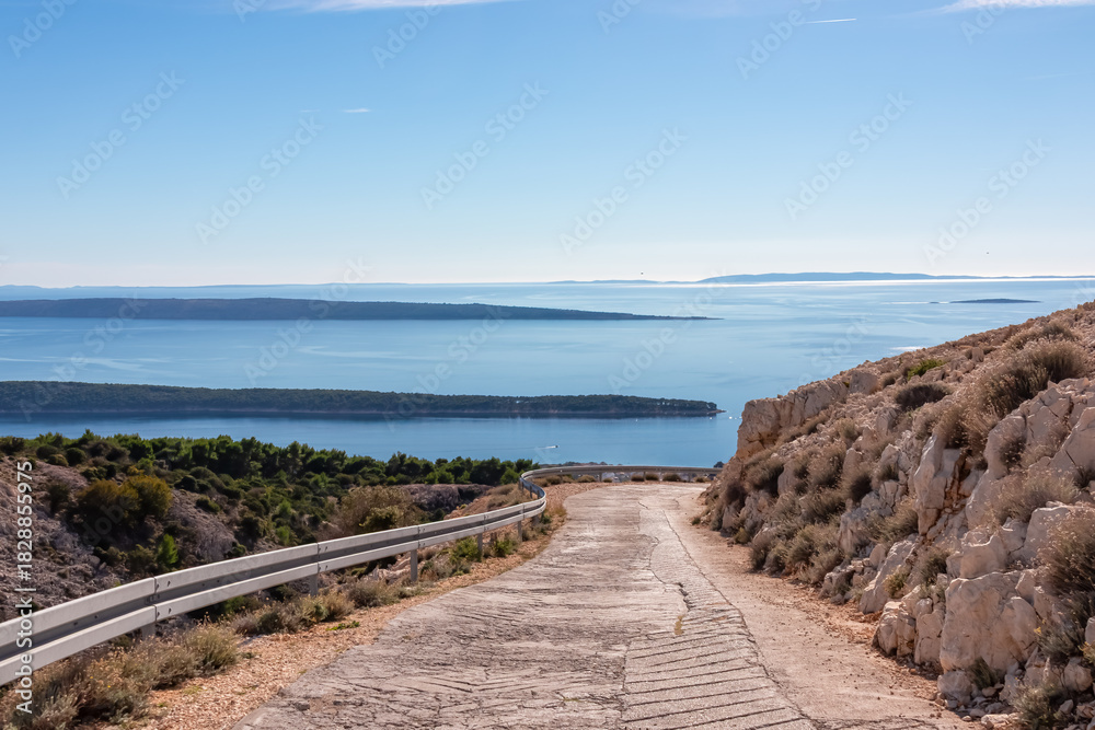 Fototapeta premium A steep concrete road descends through rocky terrain towards the Adriatic coast. This scenic route on Kamenjak mountain offers breathtaking panoramic views of the nearby islands and sea in Croatia.