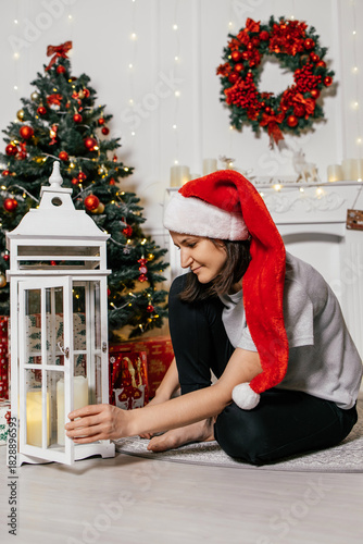 Woman wearing a red Santa hat enjoys decorating with lantern by the Christmas tree at home