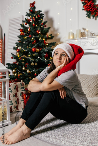 Woman in Santa hat sitting on the floor near a decorated Christmas tree with a thoughtful expression during the holiday season