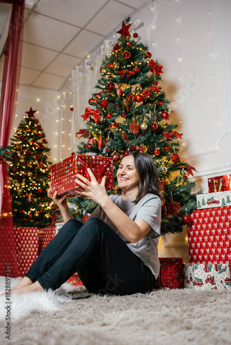 Woman joyfully opening a red gift near decorated Christmas trees in a cozy holiday setting